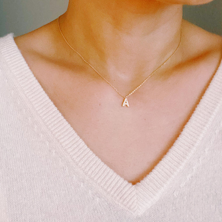 A delicate gold chain necklace with a small, pavé-set initial "A" pendant worn against a white V-neck sweater.
