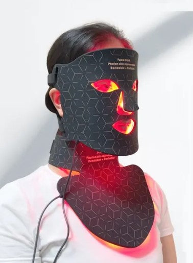 A black geometric-patterned LED therapy face and neck mask emitting red light.