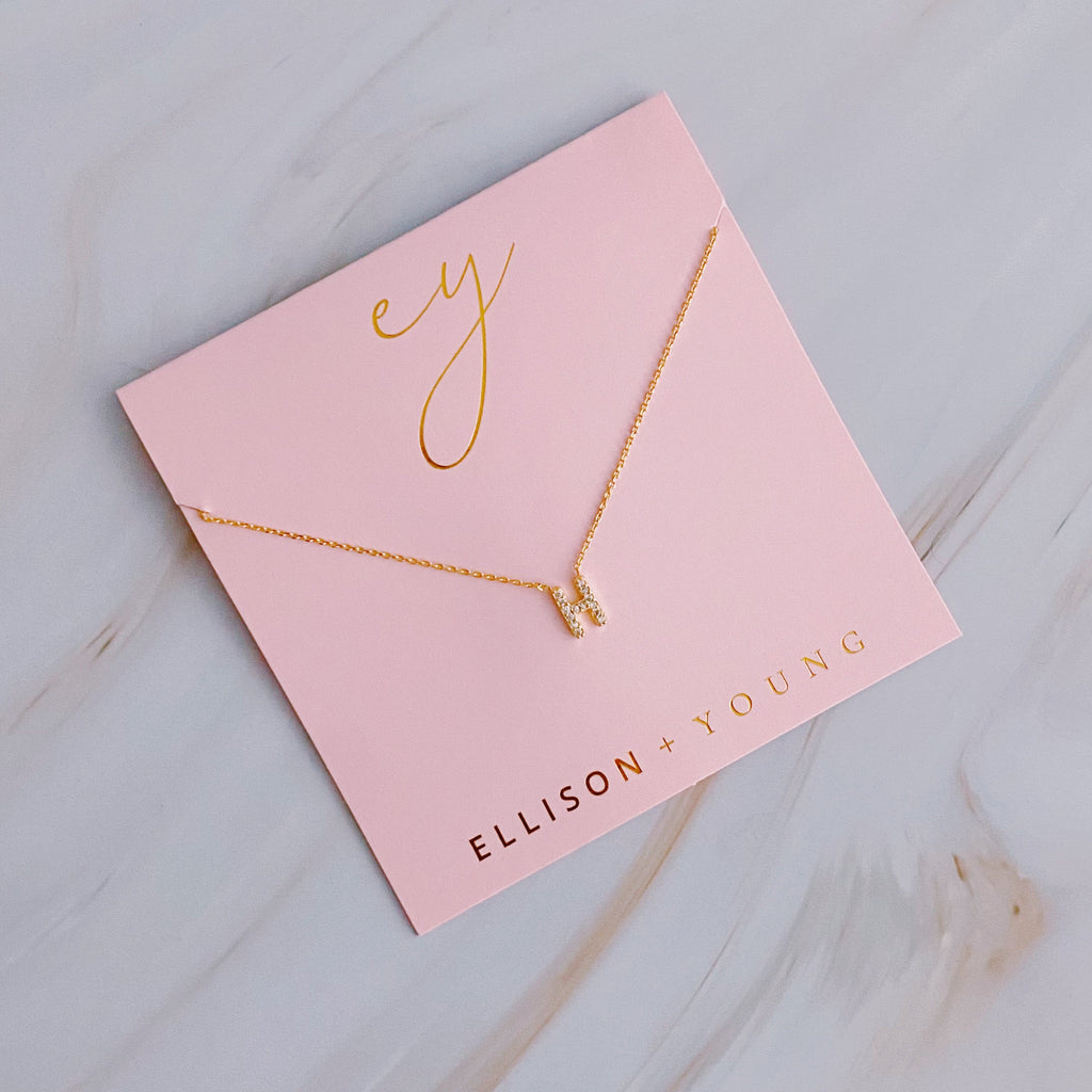A gold H initial pendant necklace on a pink card with gold lettering.