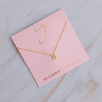 A gold H initial pendant necklace on a pink card with gold lettering.