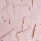 Gold initial pendant necklaces on pink card packaging with cursive gold script.