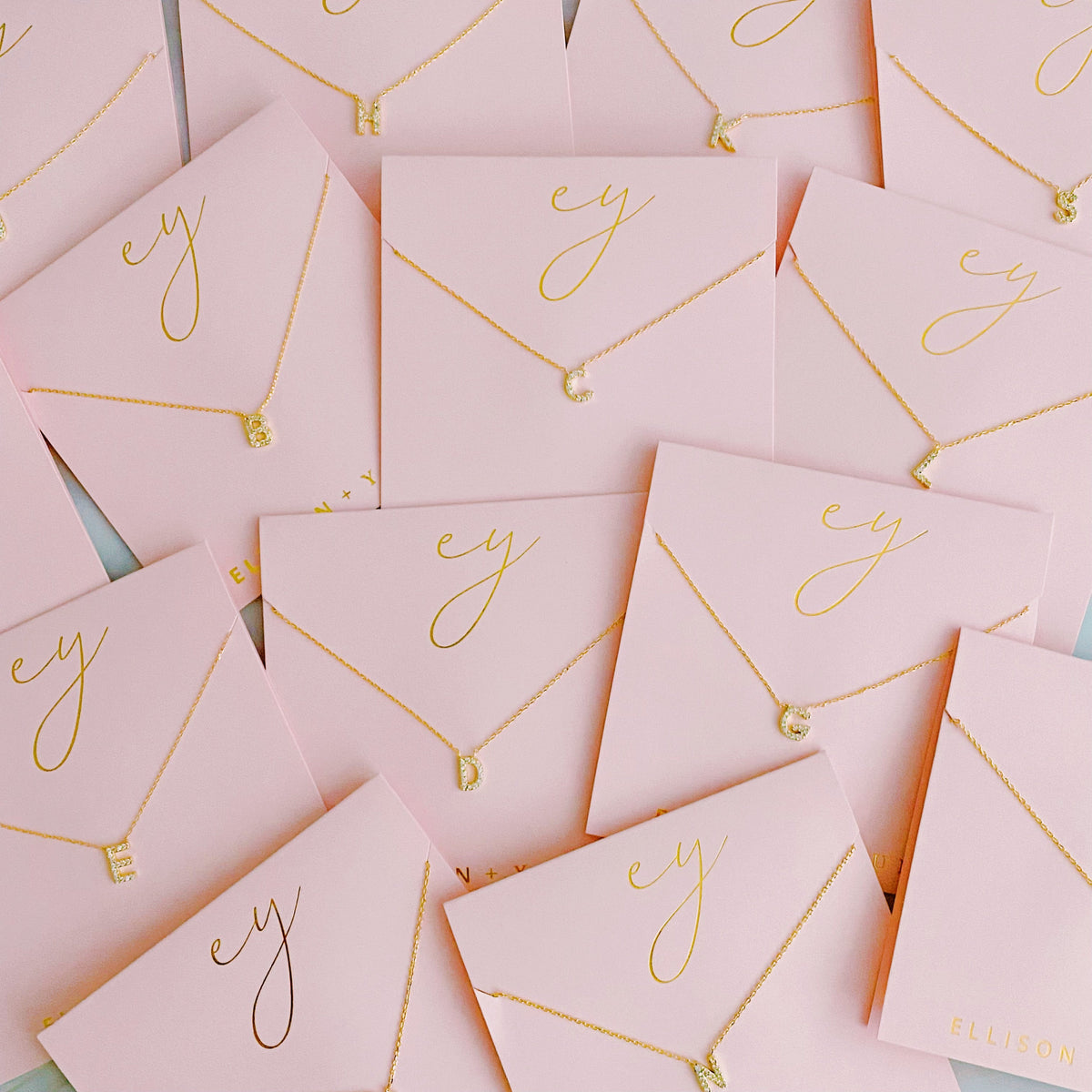 Gold initial pendant necklaces on pink card packaging with cursive gold script.