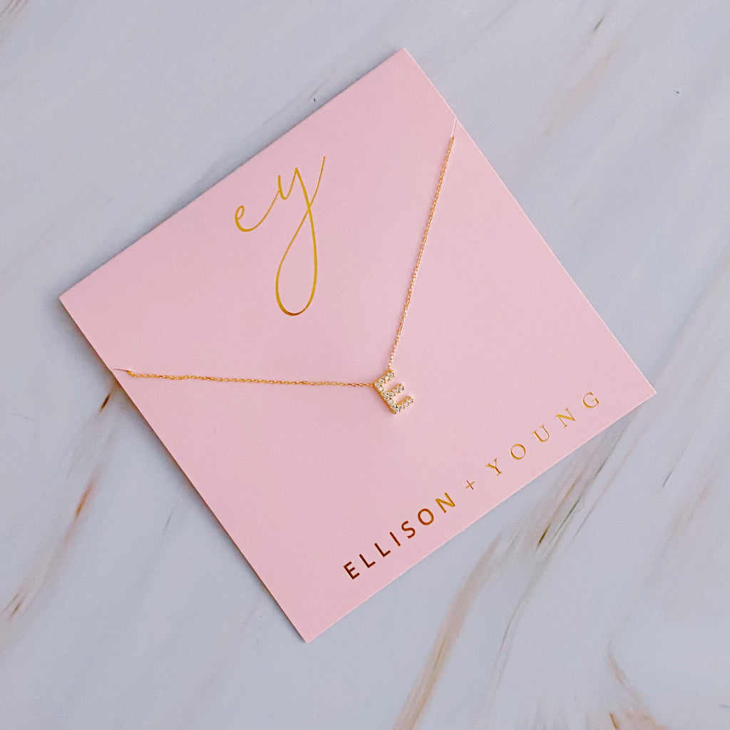 Gold letter E pendant necklace on a square pink card labeled Ellison + Young.