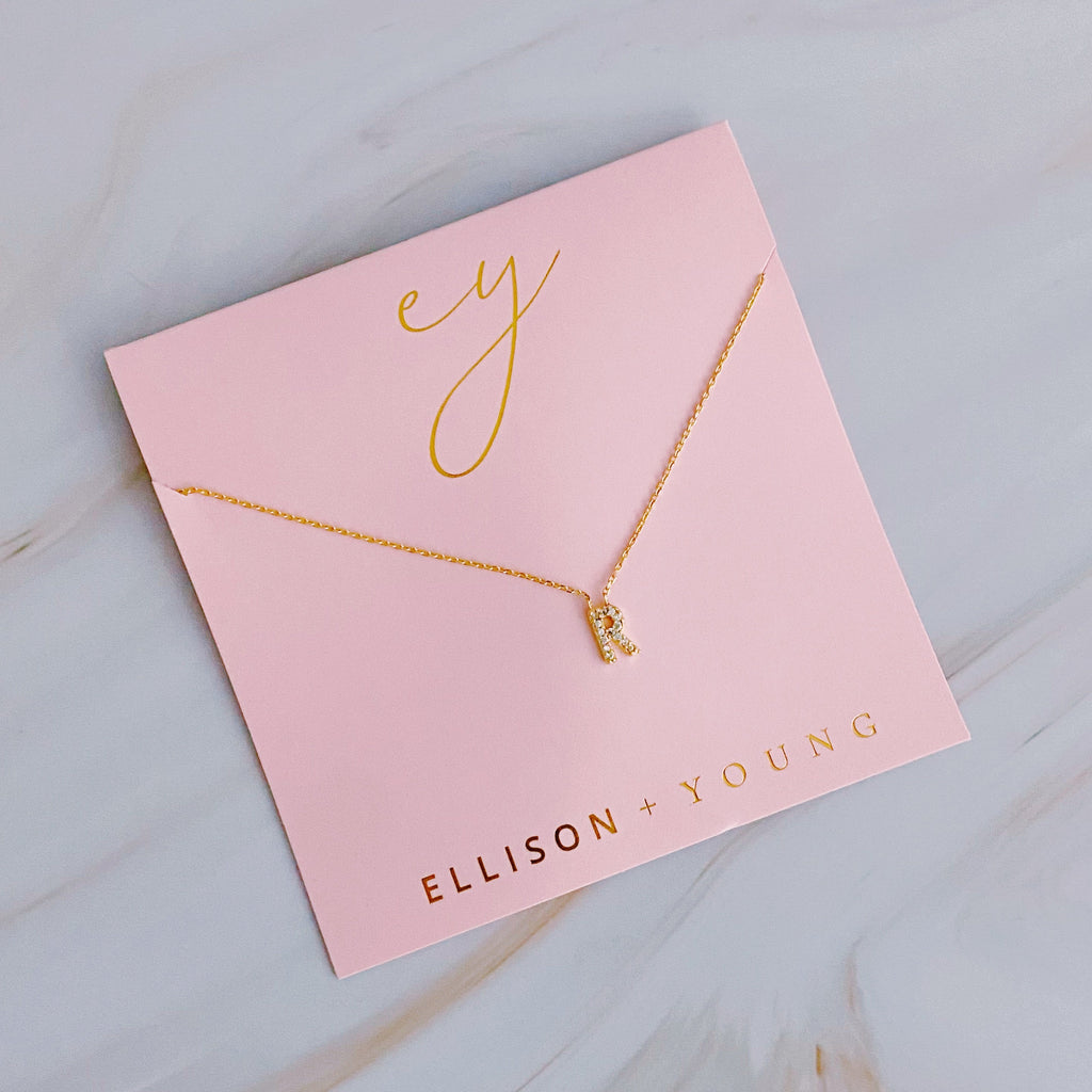 Gold initial R necklace with pavé crystals on a pink Ellison + Young card.