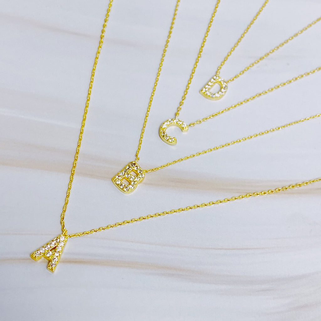 Four gold chain necklaces with crystal-paved initial pendants spelling A, B, C, and D.