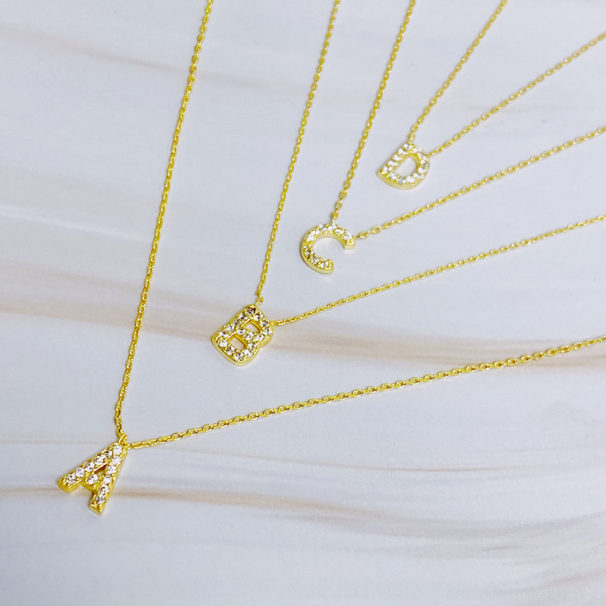 Four gold chain necklaces with crystal-paved initial pendants spelling A, B, C, and D.