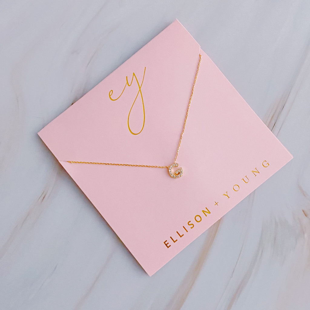 Gold initial pendant necklace with crystal accents displayed on pink Ellison + Young packaging.