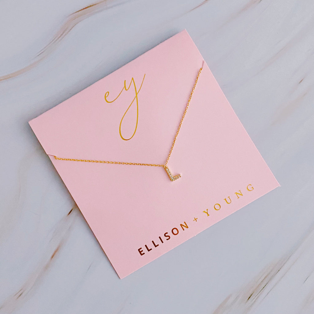 A gold initial "L" pendant necklace with crystals, displayed on a pink Ellison + Young card.