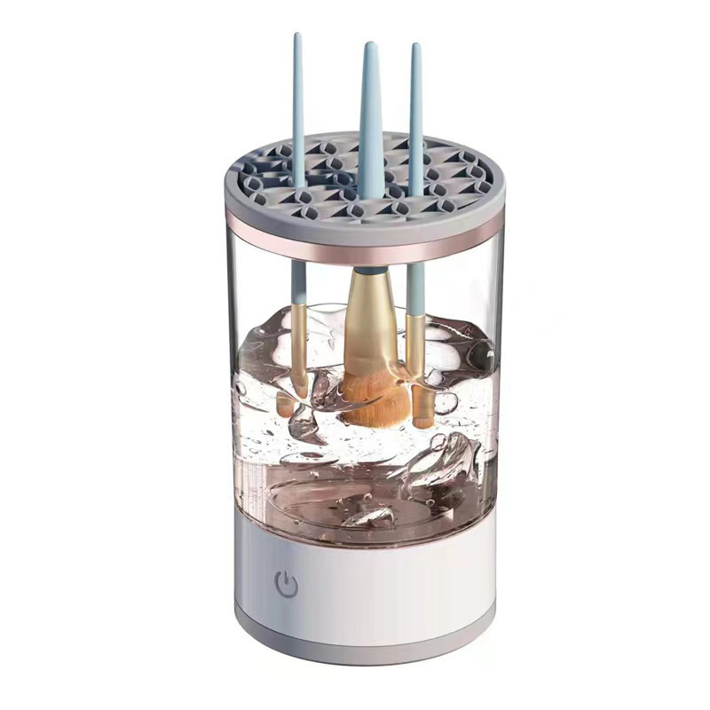 A clear, cylindrical makeup brush cleaner holding four brushes submerged in swirling water above a white base.