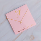 A gold initial "K" pendant necklace displayed on branded pink packaging.