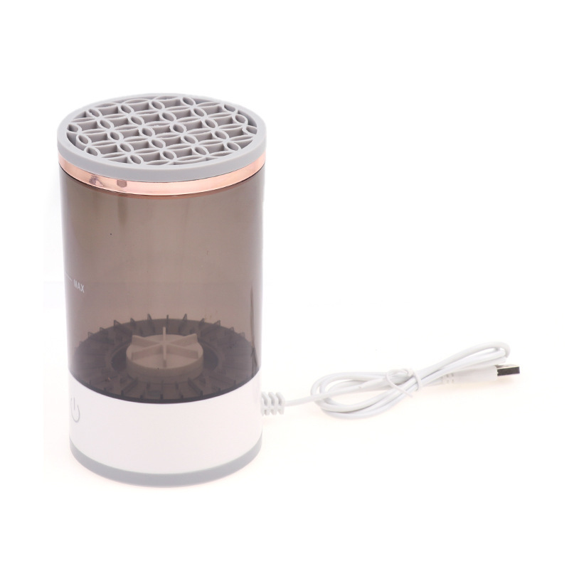 A cylindrical makeup brush cleaner with a white base, smoky grey container, rose gold accent, and a USB cord.