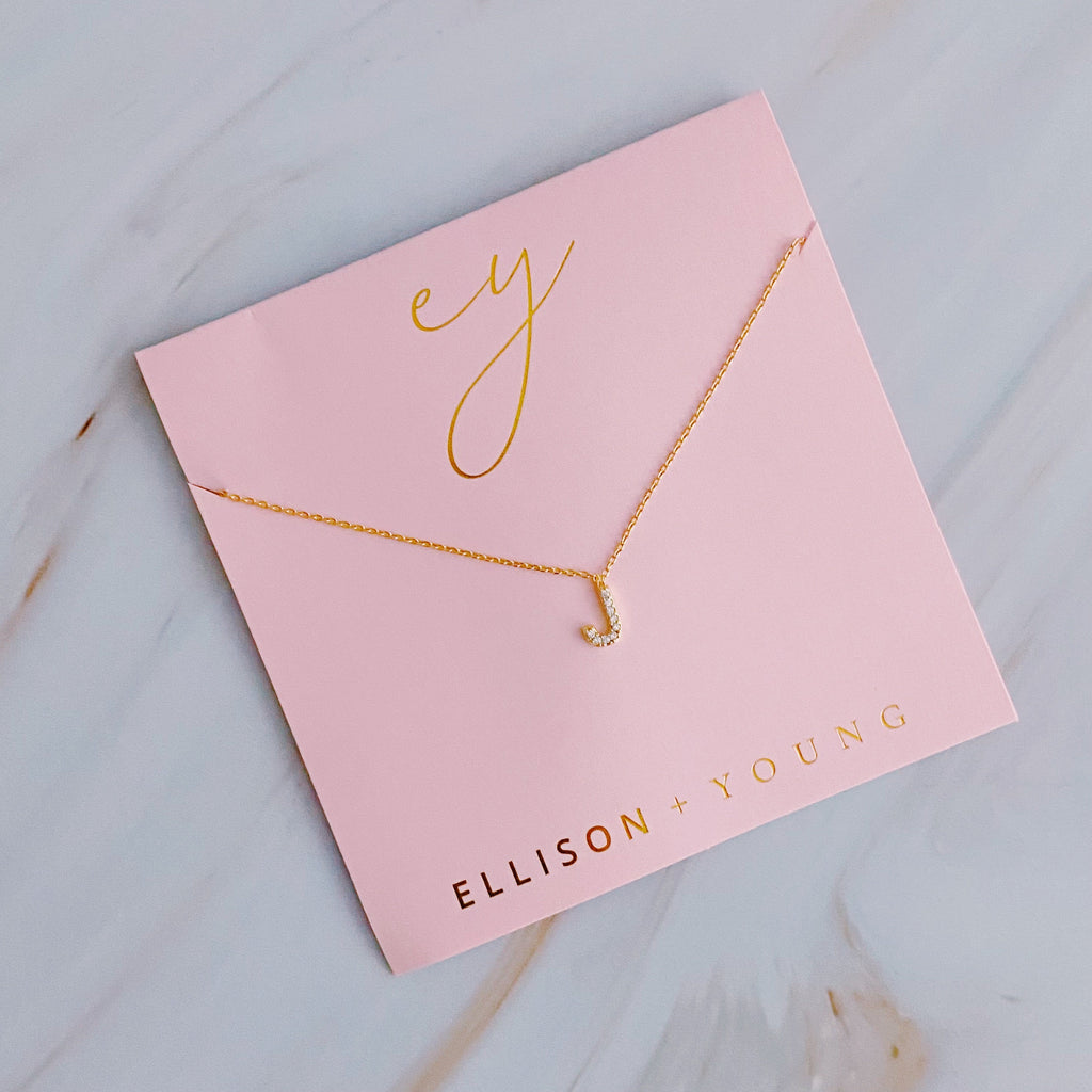 Gold initial J pendant necklace with tiny gems, displayed on a pink card with gold lettering.