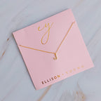 Gold initial J pendant necklace with tiny gems, displayed on a pink card with gold lettering.