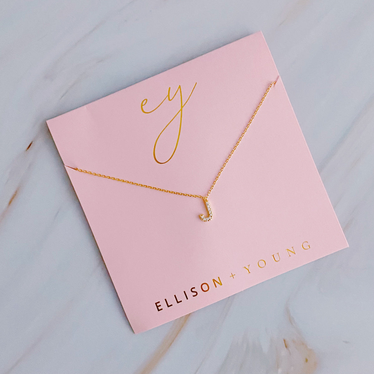 Gold initial J pendant necklace with tiny gems, displayed on a pink card with gold lettering.