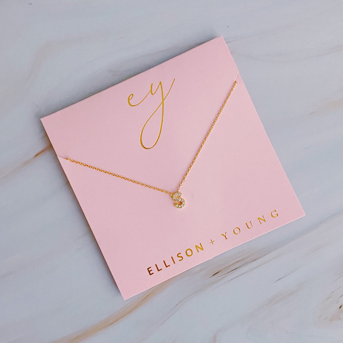 A gold chain necklace with a crystal-embellished letter S pendant on pink packaging.