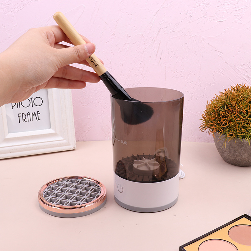 A hand dips a makeup brush into a translucent dark-brown and white electric makeup brush cleaner.