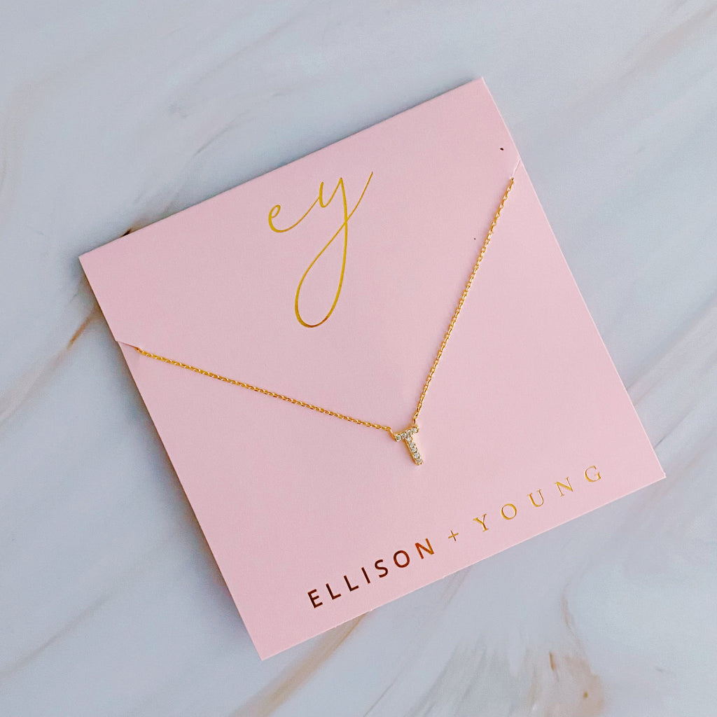 A gold initial necklace with a "T" pendant on a pink backing card labeled "ELLISON + YOUNG".