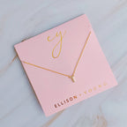 A gold initial necklace with a "T" pendant on a pink backing card labeled "ELLISON + YOUNG".