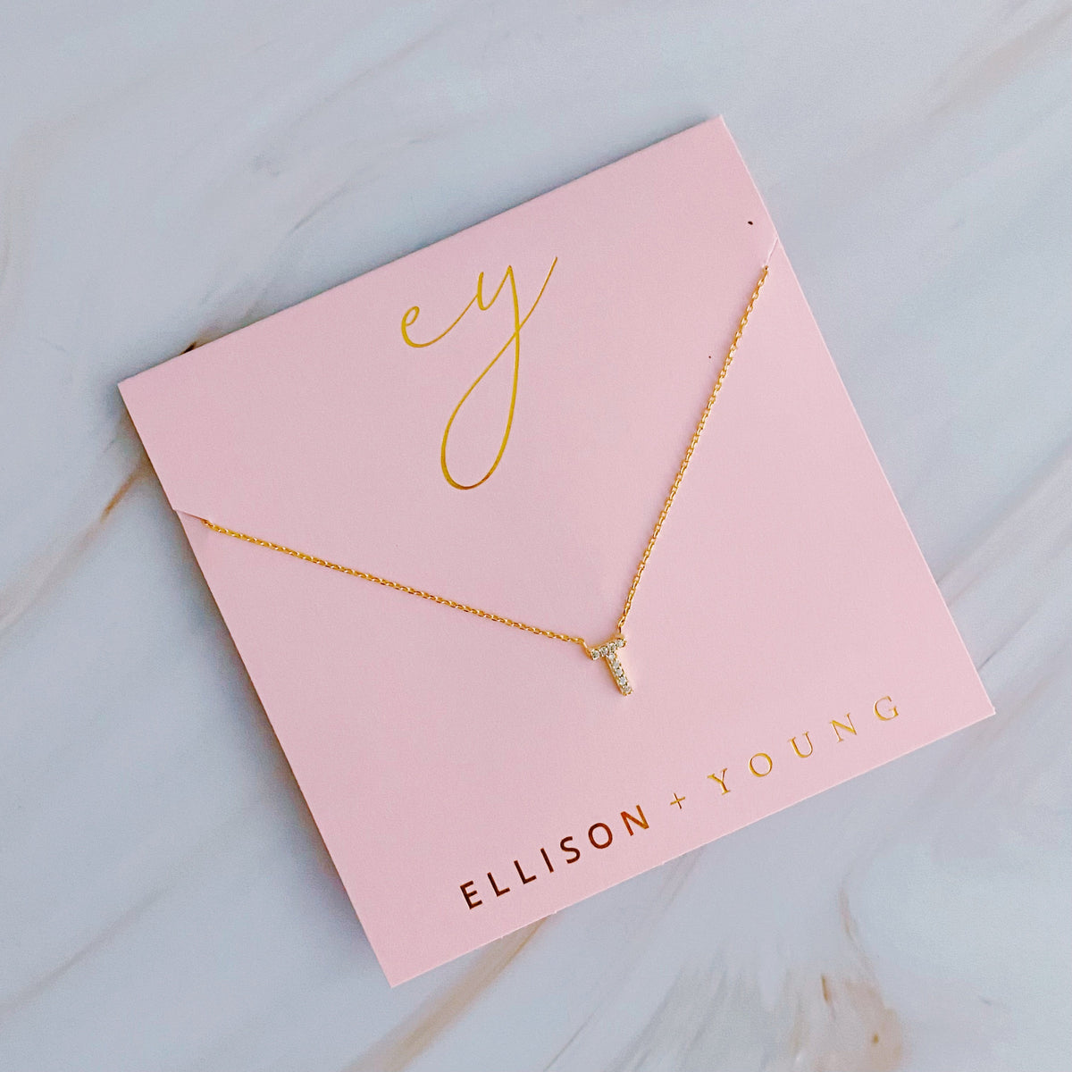 A gold initial necklace with a "T" pendant on a pink backing card labeled "ELLISON + YOUNG".
