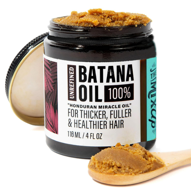 Open jar of brown, grainy batana hair oil with a wooden spoon holding a scoop in front.