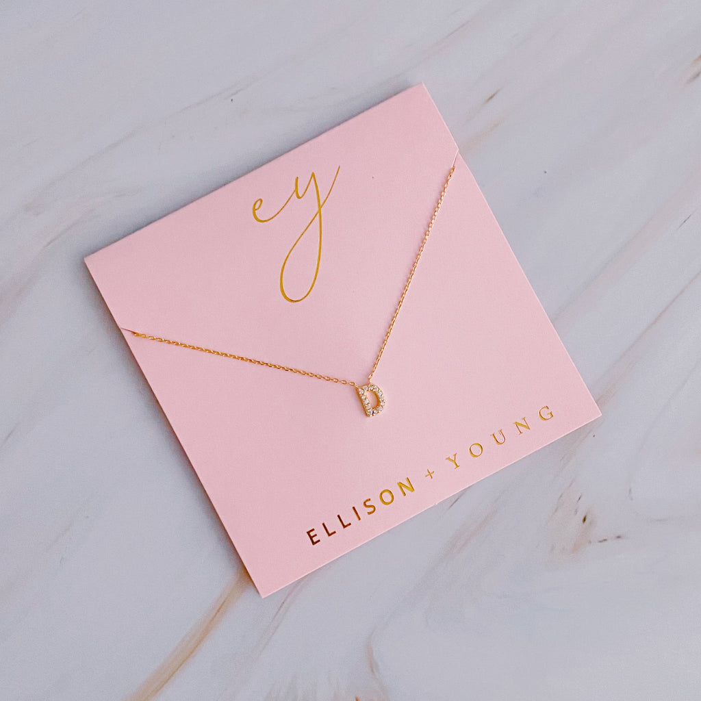 A gold initial D pendant necklace displayed on a pink packaging card.