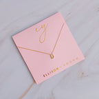 A gold initial D pendant necklace displayed on a pink packaging card.