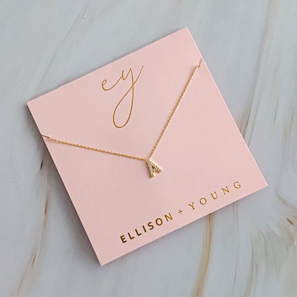 Gold initial pendant necklace featuring letter A on pink packaging branded Ellison + Young.