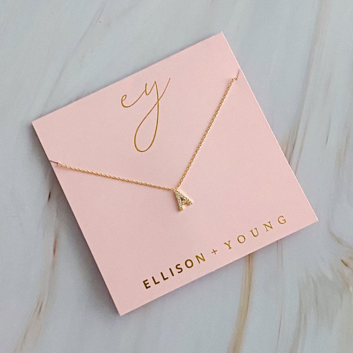 Gold initial A pendant necklace on a square pink display card labeled Ellison + Young.